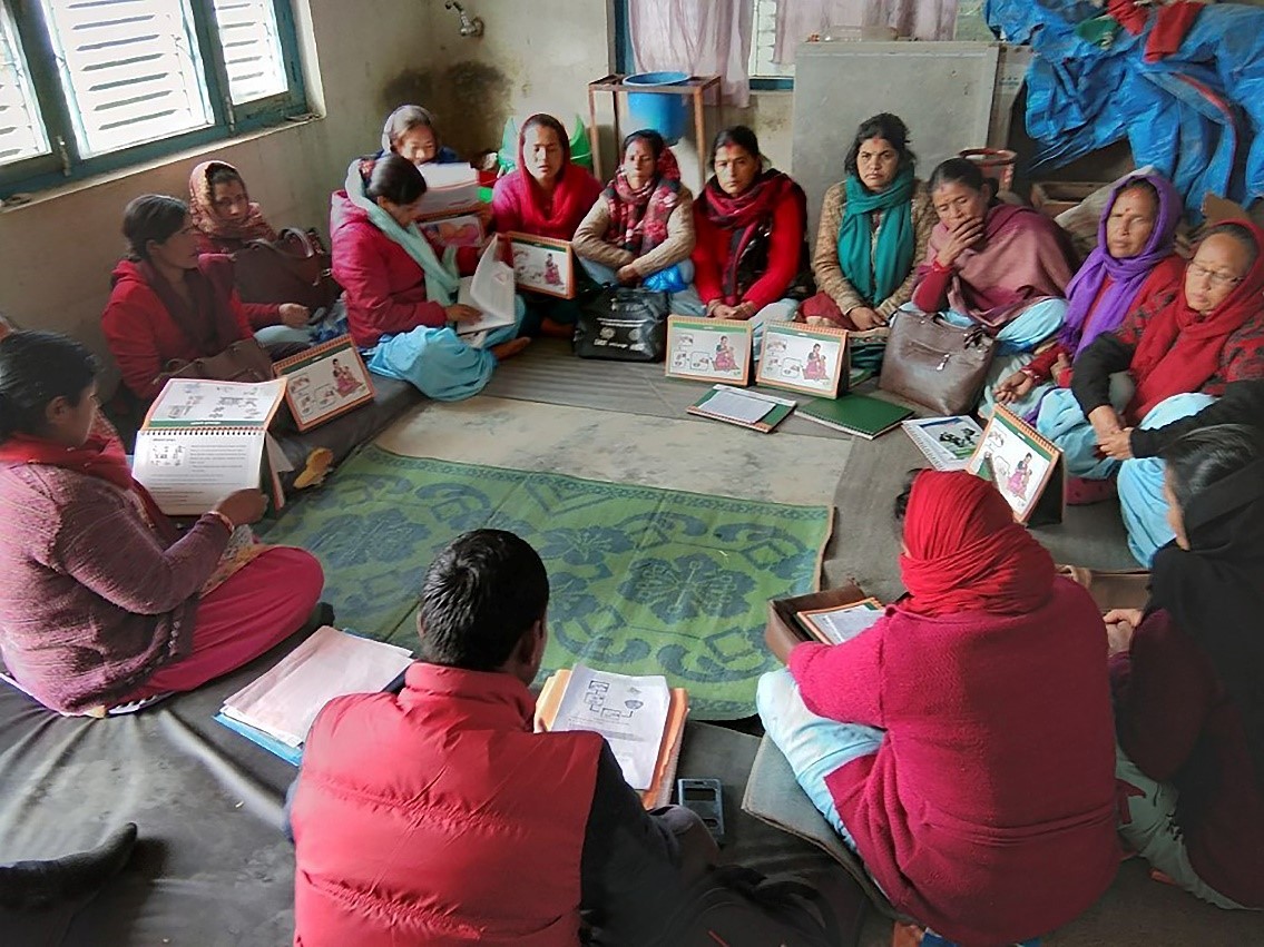 A group of 14 Nepalese women and 1 man sit in a circle on a green carpet. Most are wearing red and headscarves.