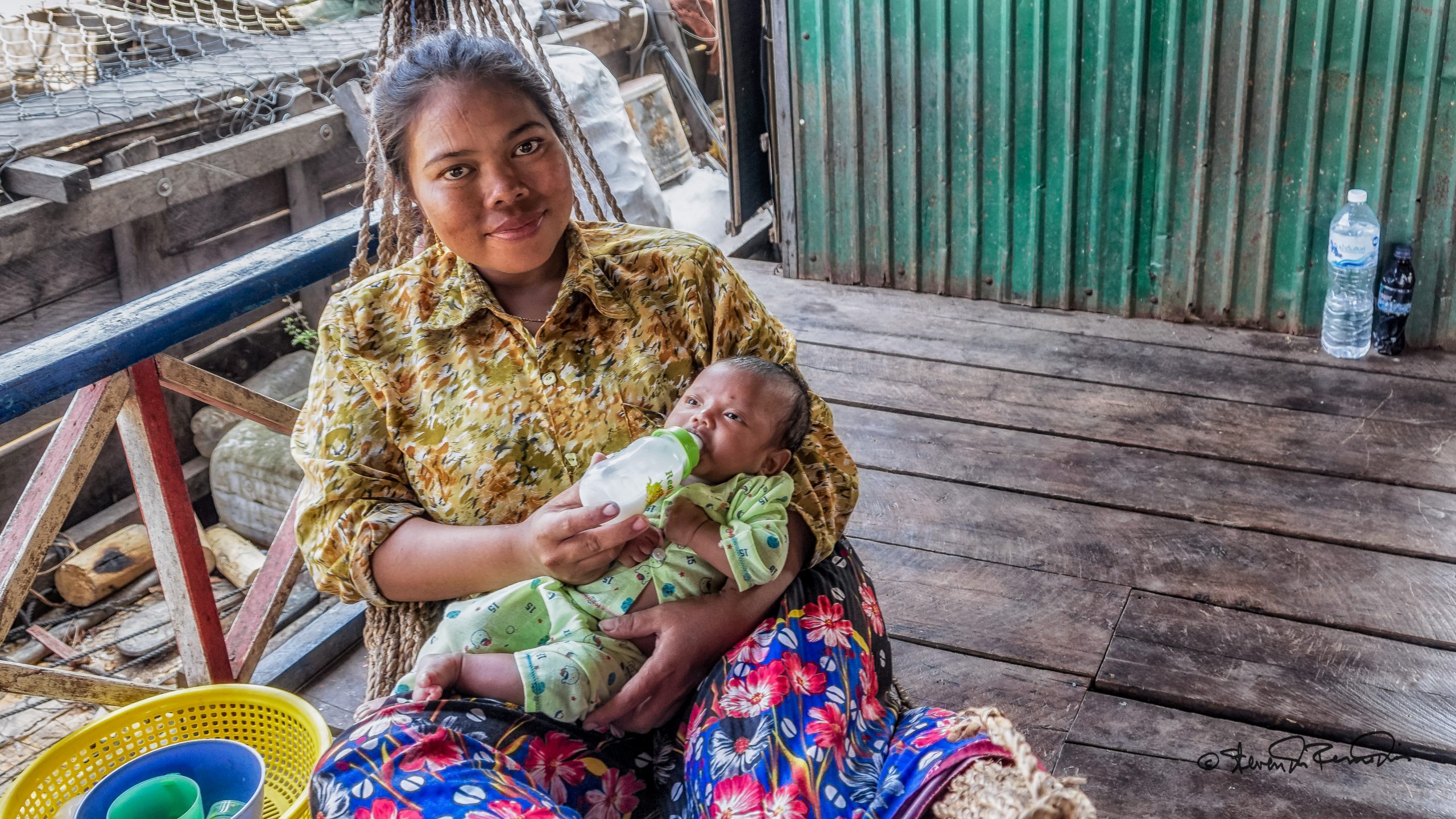 Cambodian woman bottle feeds a baby while sitting in a wooden structure near a boat - she's looking at the camera and smiling