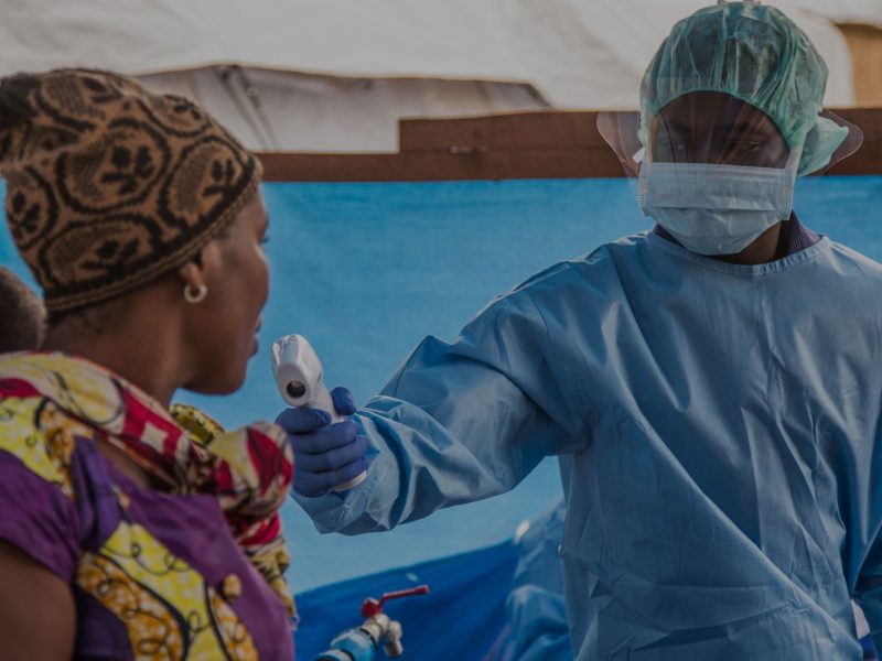 African man in PPE takes the temperature of a woman with a baby on her back