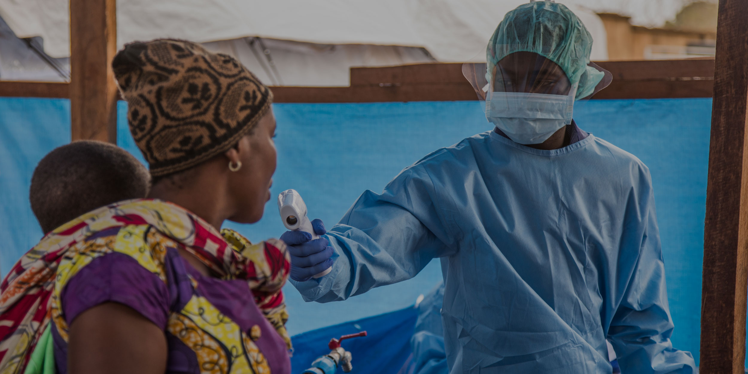 African man in PPE takes the temperature of a woman with a baby on her back