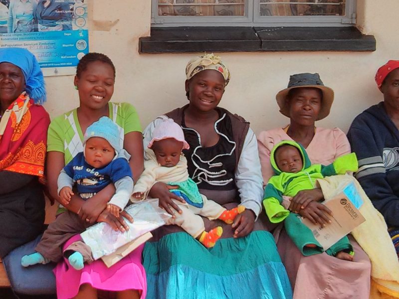 Five women sit on a bench outside a ‘Treatment room’ and ‘Consultation room’. They are brightly clothed, smiling and holding three babies.