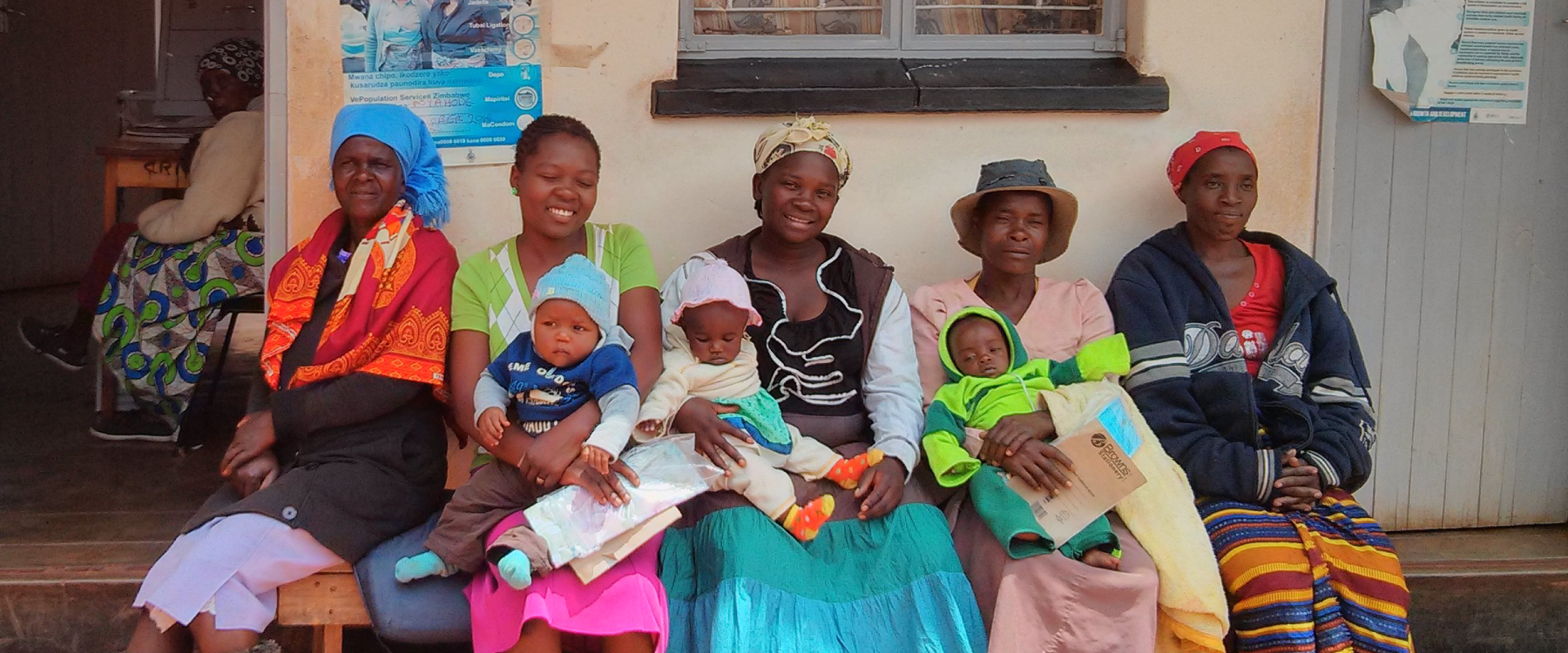 Five women sit on a bench outside a ‘Treatment room’ and ‘Consultation room’. They are brightly clothed, smiling and holding three babies.