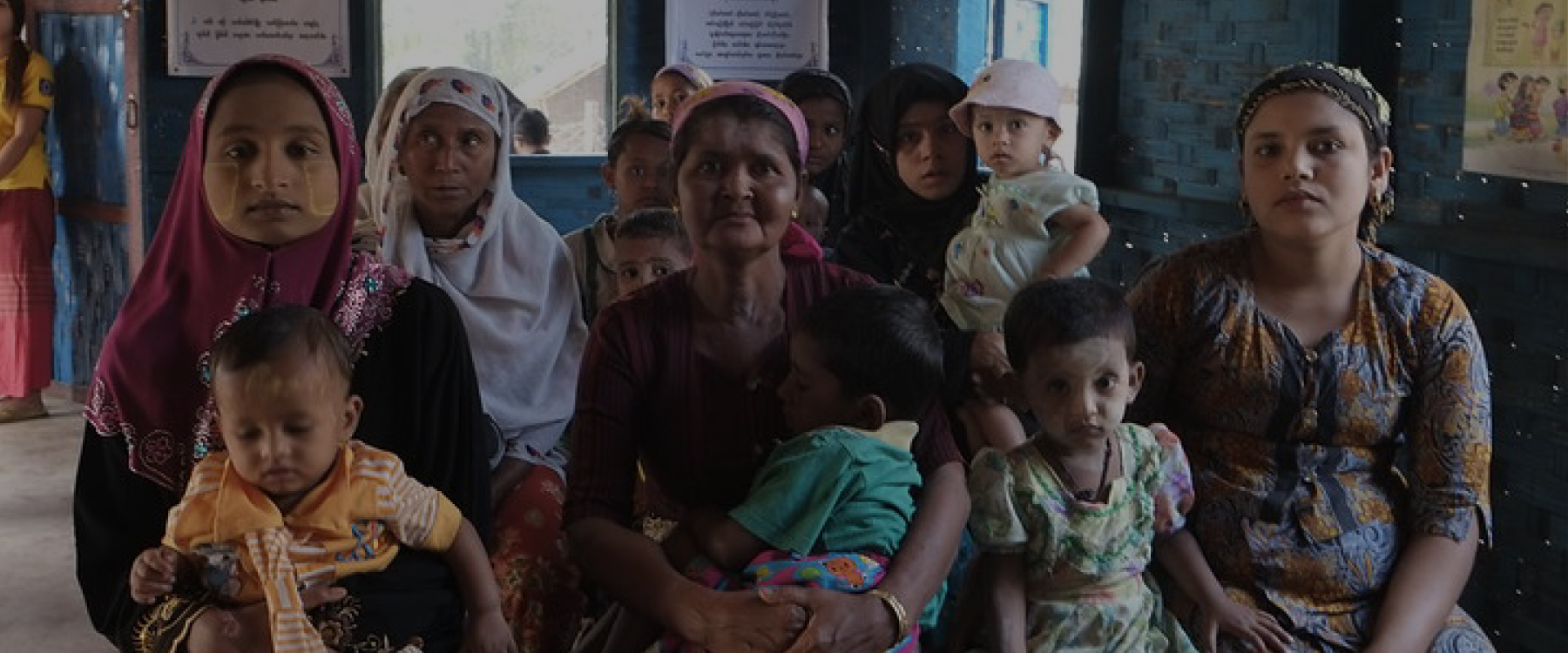 Seated women wearing head scarves and with children on their knees stare unsmiling at the camera. They are in a waiting room.