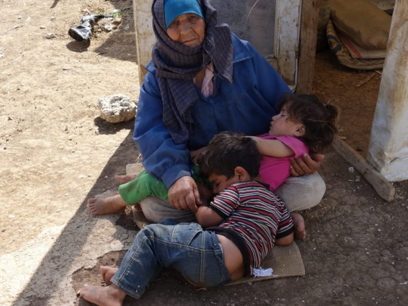 Old woman in blue headscarf sitting on the ground cradling a sleeping girl and boy