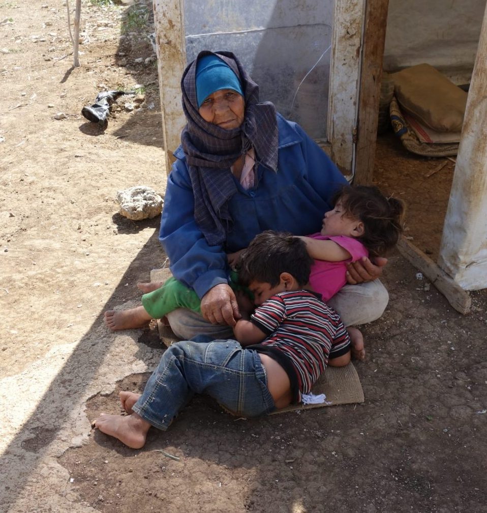 Old woman in blue headscarf sitting on the ground cradling a sleeping girl and boy