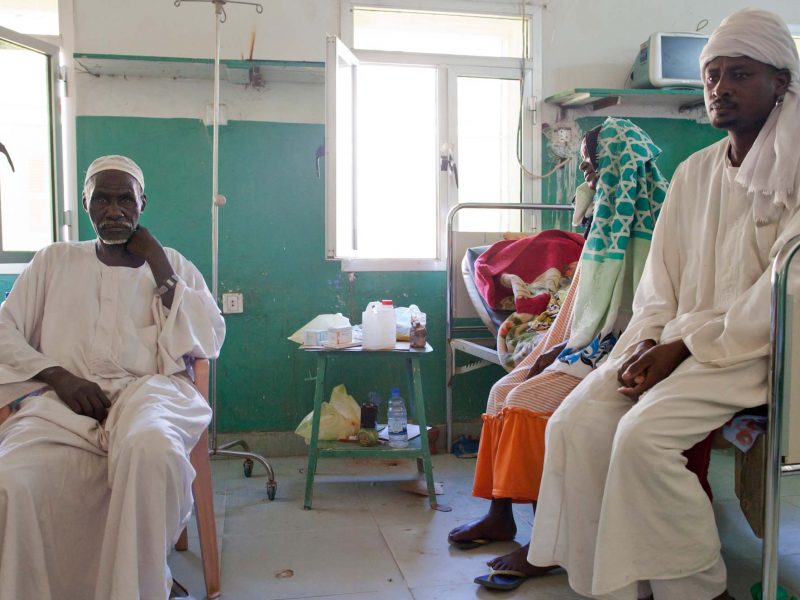 An African women and man sit on a hospital bed while a man sits on a chair, staring at the camera