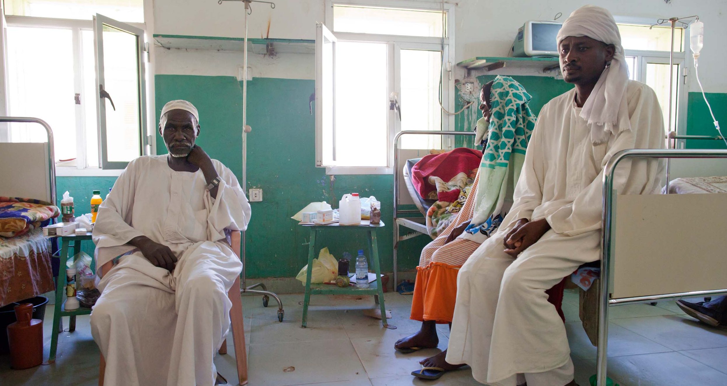 An African women and man sit on a hospital bed while a man sits on a chair, staring at the camera