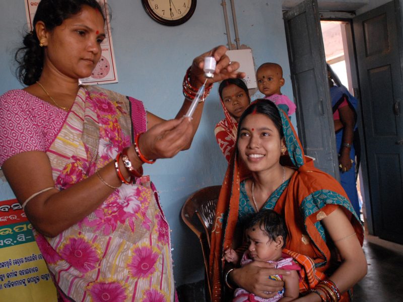 Standing Indian woman files a syringe while a seated mother and her baby look on