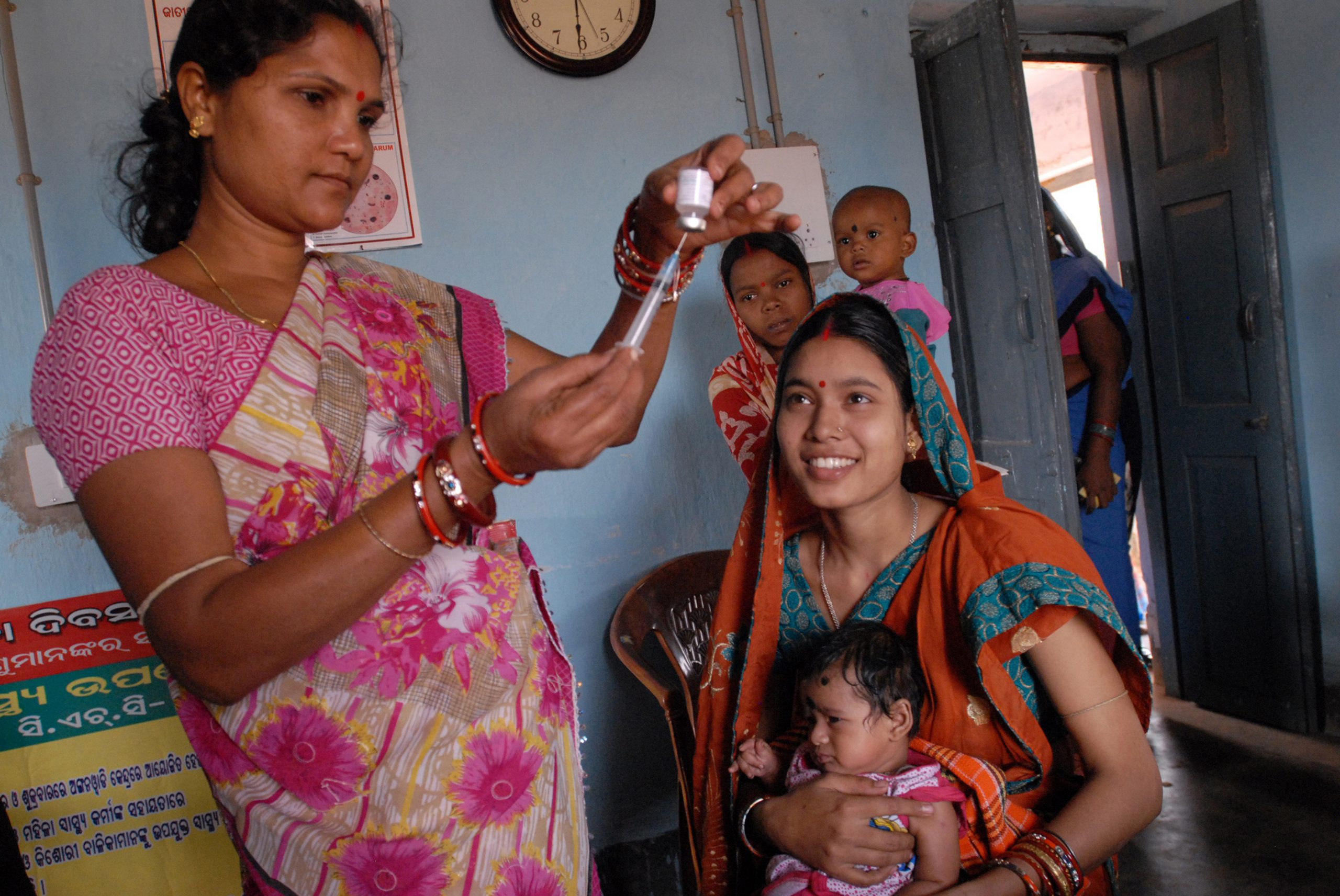 Standing Indian woman files a syringe while a seated mother and her baby look on