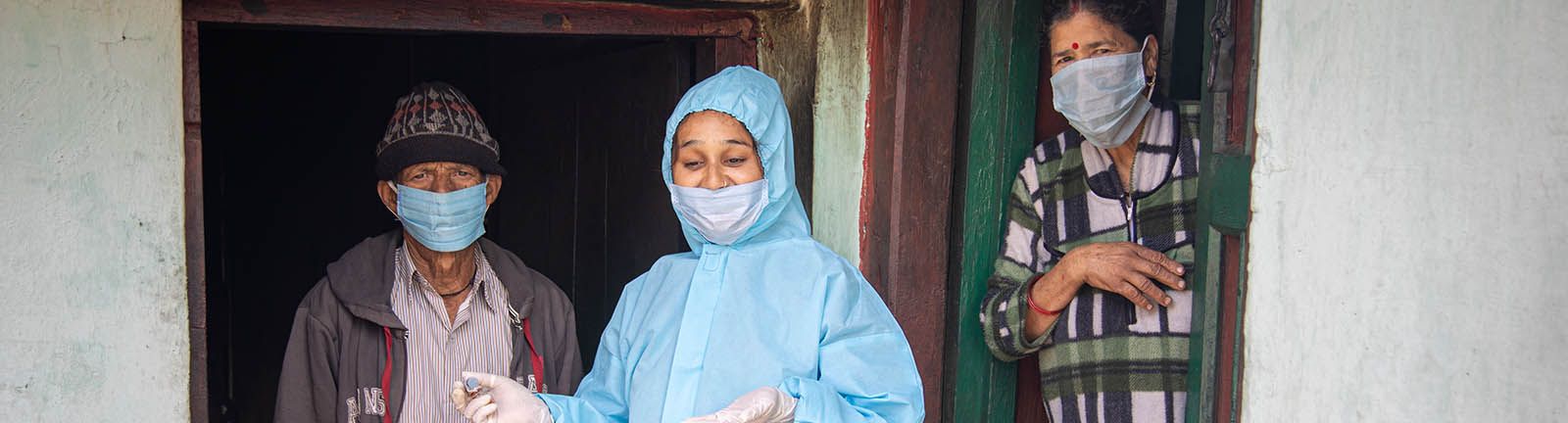 An India woman in blue PPE stands between a man and a woman both wearing face masks