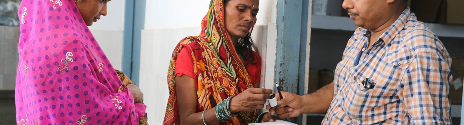 Indian woman in an orange sari takes a small medicine bottle from a man while a woman in a pink sari looks on