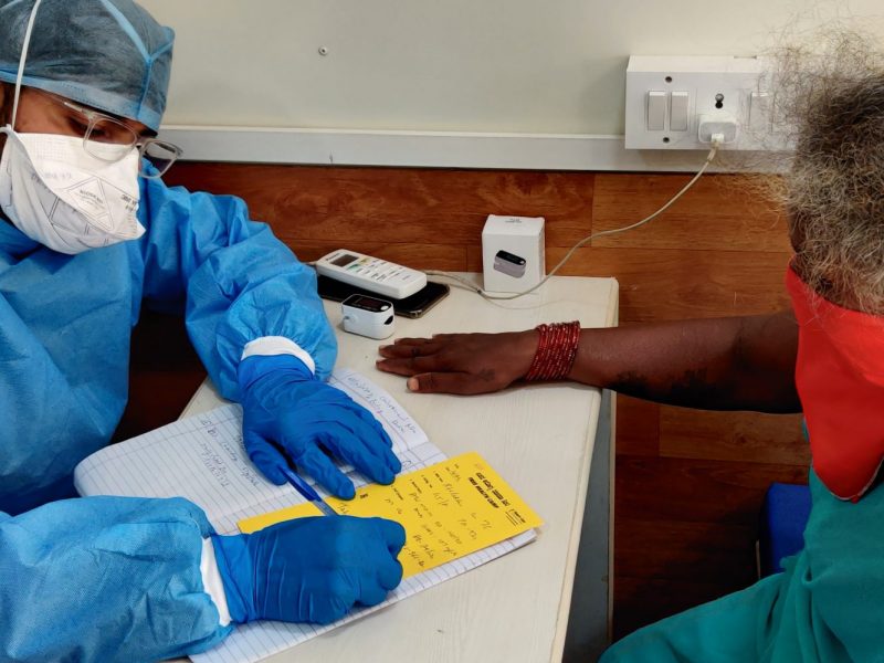 A male Indian doctor wearing PPE sits at a tale writing - sitting opposite is a older Indian lady wearing an orange mask