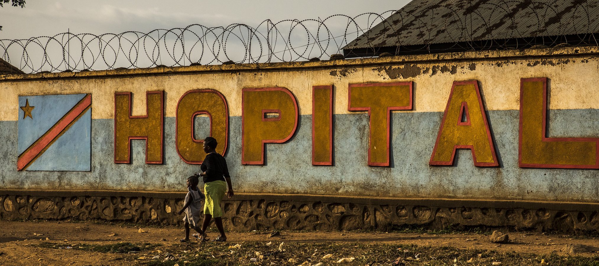 An African woman and small child walk past a wall with the word 'HOPITAL' and barbed wire across the top