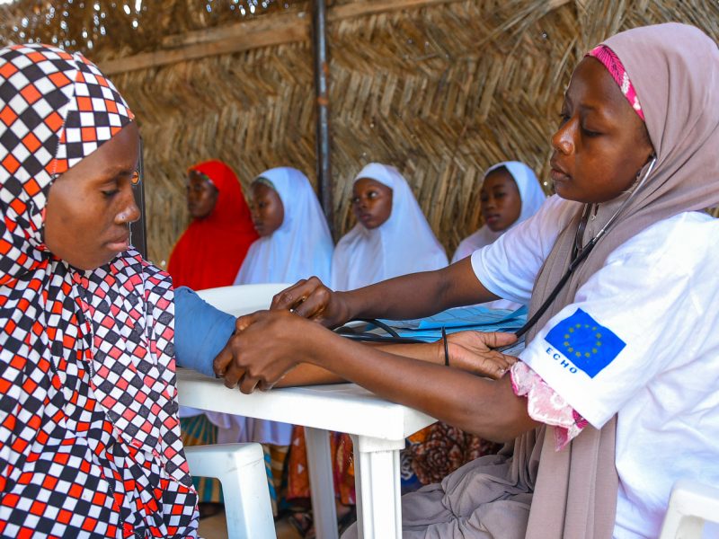 An African nurse sits tending to another African woman while three women sit in the background, all beneath a wicker shelter