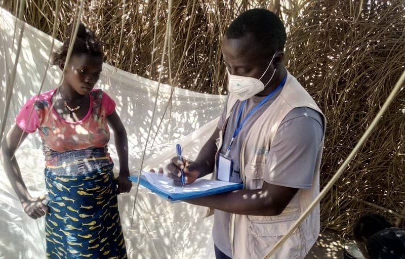 African man wearing a face mask writes on a clipboard while a pregnant Africa woman watches on