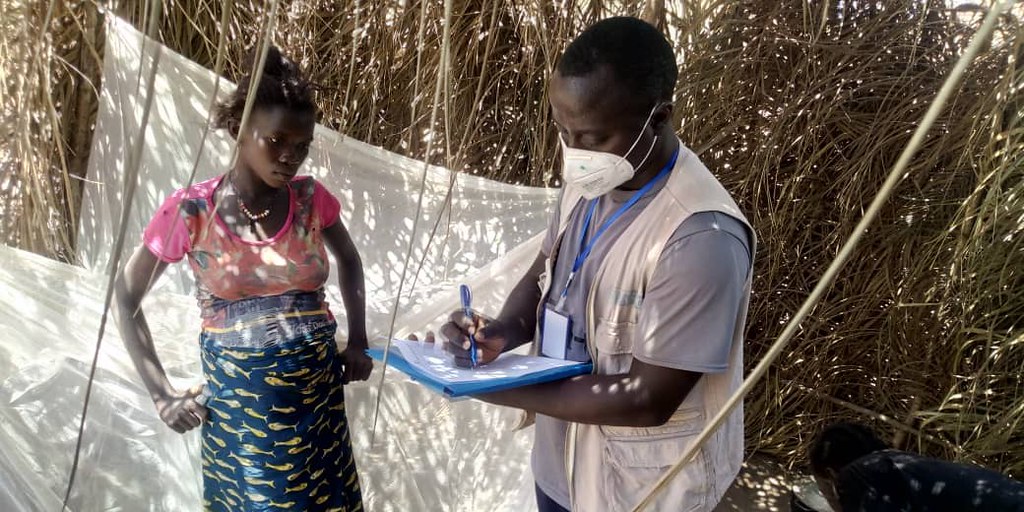 African man wearing a face mask writes on a clipboard while a pregnant Africa woman watches on