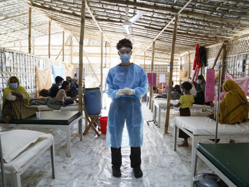 A woman in blue scrubs stands looking at the viewer in a hut with women and children sitting on beds