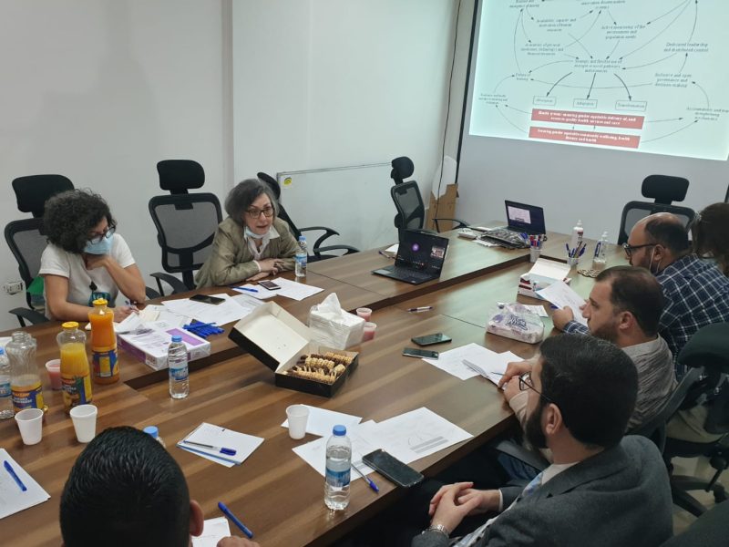 Men and women sitting around a table, talking, with a complex diagram is projected onto the wall beside them