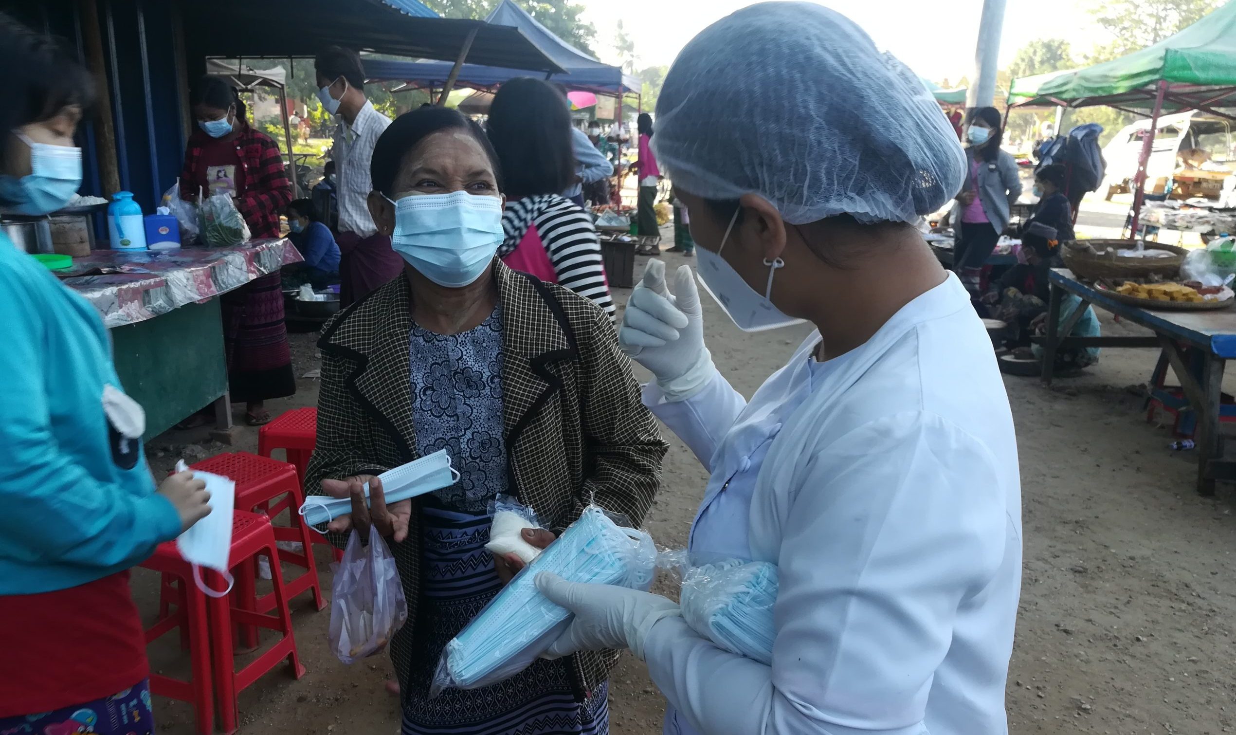 Woman in face mask and hairnet hands out masks to another masked lady in a market
