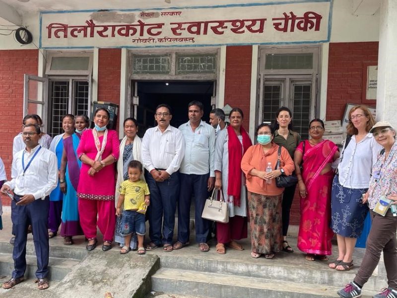 A row of standing smiling women and men outside a health facility - some of the women are wearing red saris