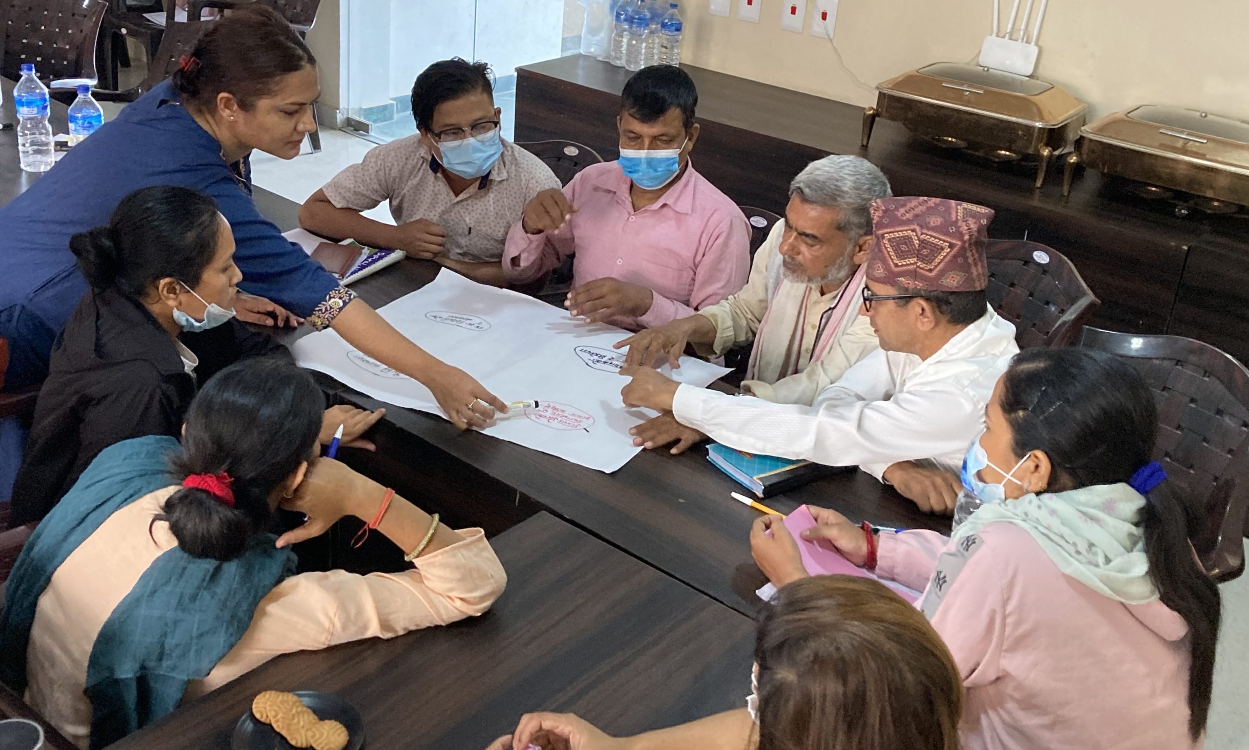 A group of seated Nepali men and women look at a sheet of flipcahrt paper while a standing women in a blue dress points at the sheet