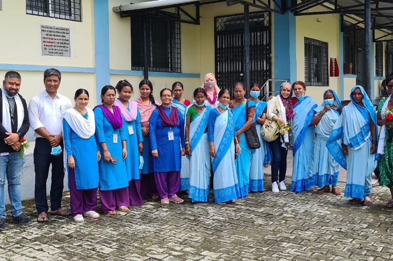 A row of smiling, standing women and a few men outside a health centre. Many of the women are wearing blue saris