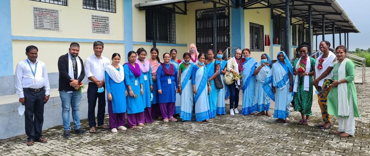 A row of smiling, standing women and a few men outside a health centre. Many of the women are wearing blue saris