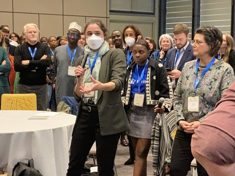 People standing in a large room listening to a standing masked woman speak into a microphone