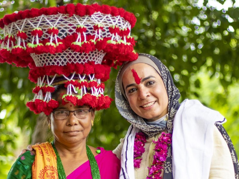 Two standing, smiling women - one is Nepali and wears a very large tassled hat, the other a headscarf and a red stripe down the middle of her forehead