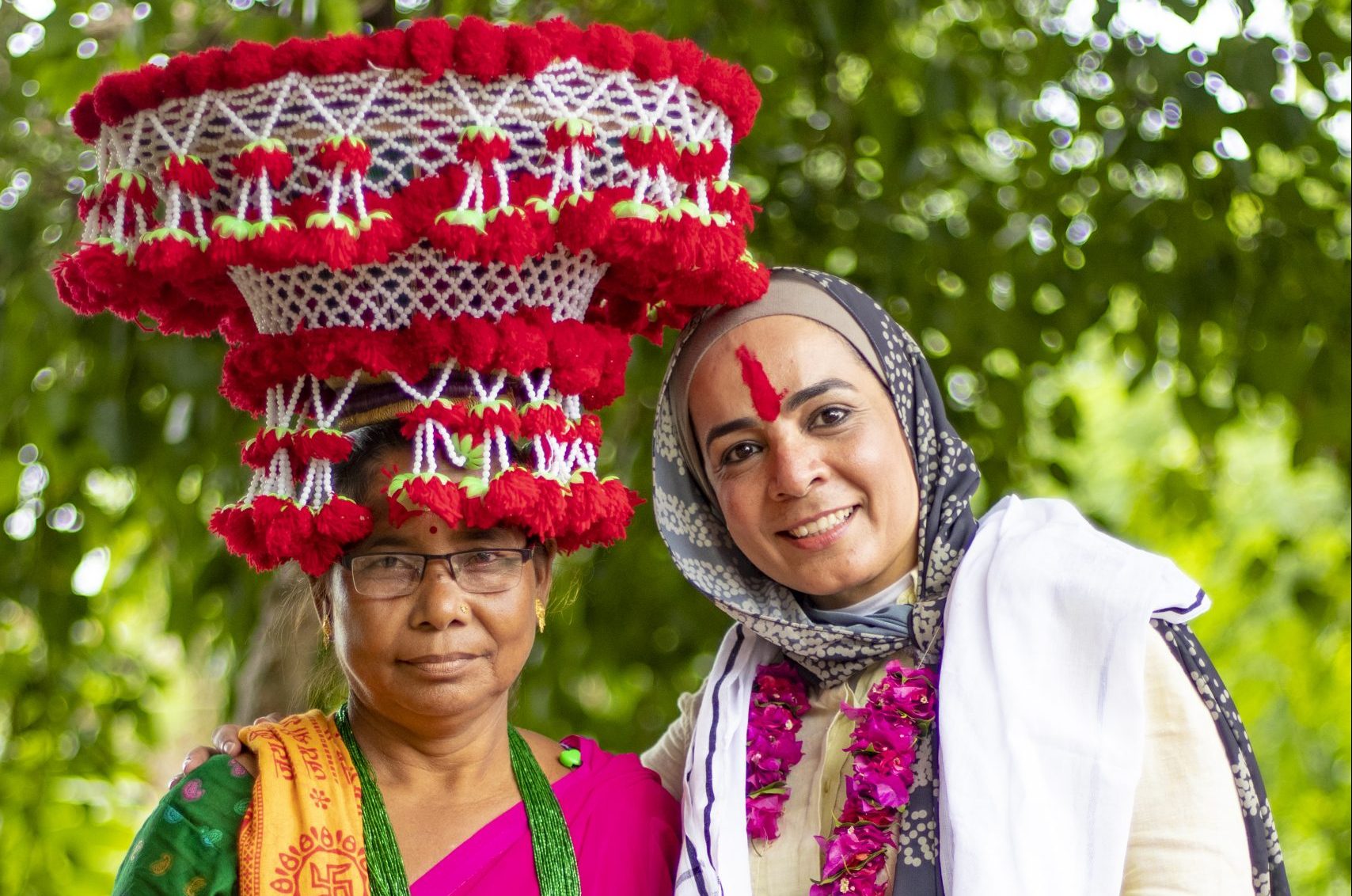 Two standing, smiling women - one is Nepali and wears a very large tassled hat, the other a headscarf and a red stripe down the middle of her forehead