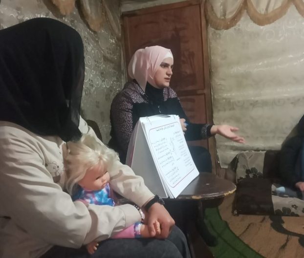 A woman wearing a pink headscarf talks to other women (out of shot) while another woman sits nearby holding a doll on her knee. There is a large diagram on display as well.