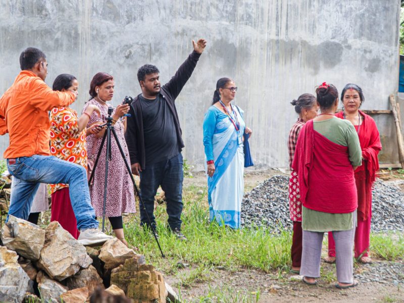 A Nepali man in black stands with his arm raised to some in the distance while a group of female health workers stand around a camera on a tripod