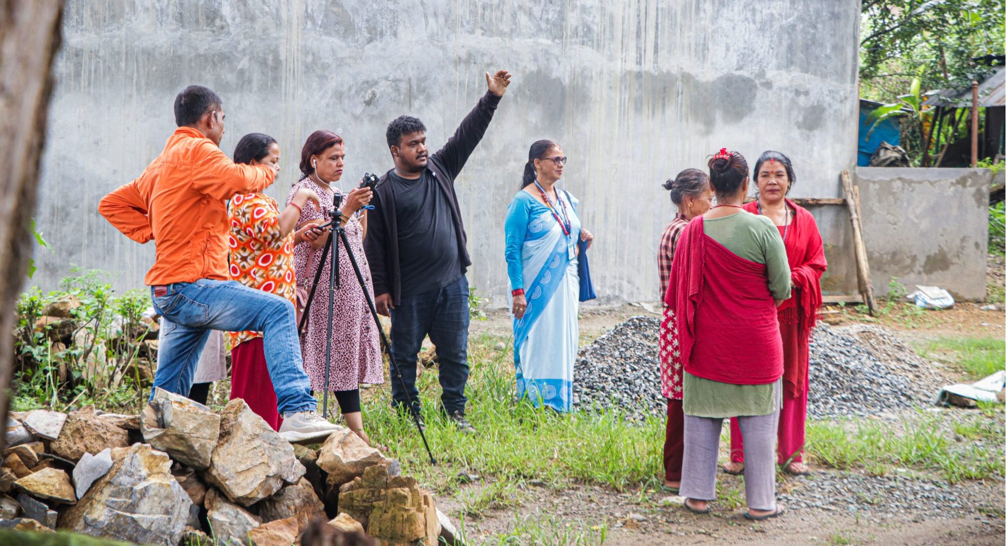 A Nepali man in black stands with his arm raised to some in the distance while a group of female health workers stand around a camera on a tripod