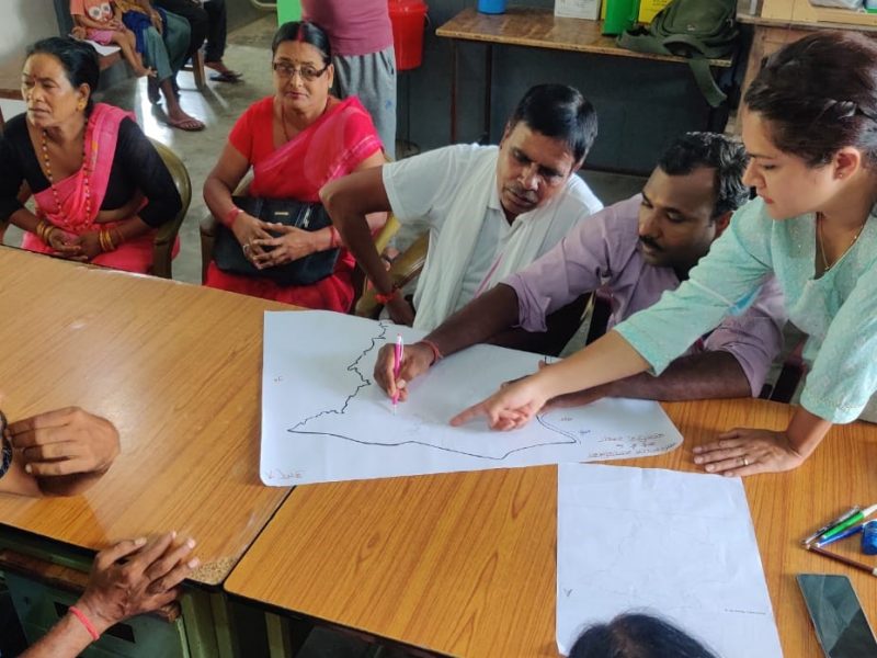 A standing woman in green talking to a group of men and women seated around a diagram they are drawing