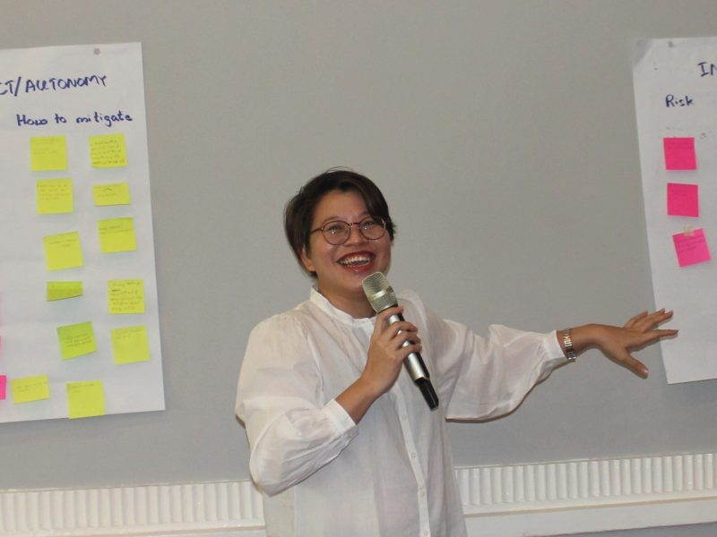 A smiling Burmese woman stands pointing to post-its on a wall while speaking into a microphone