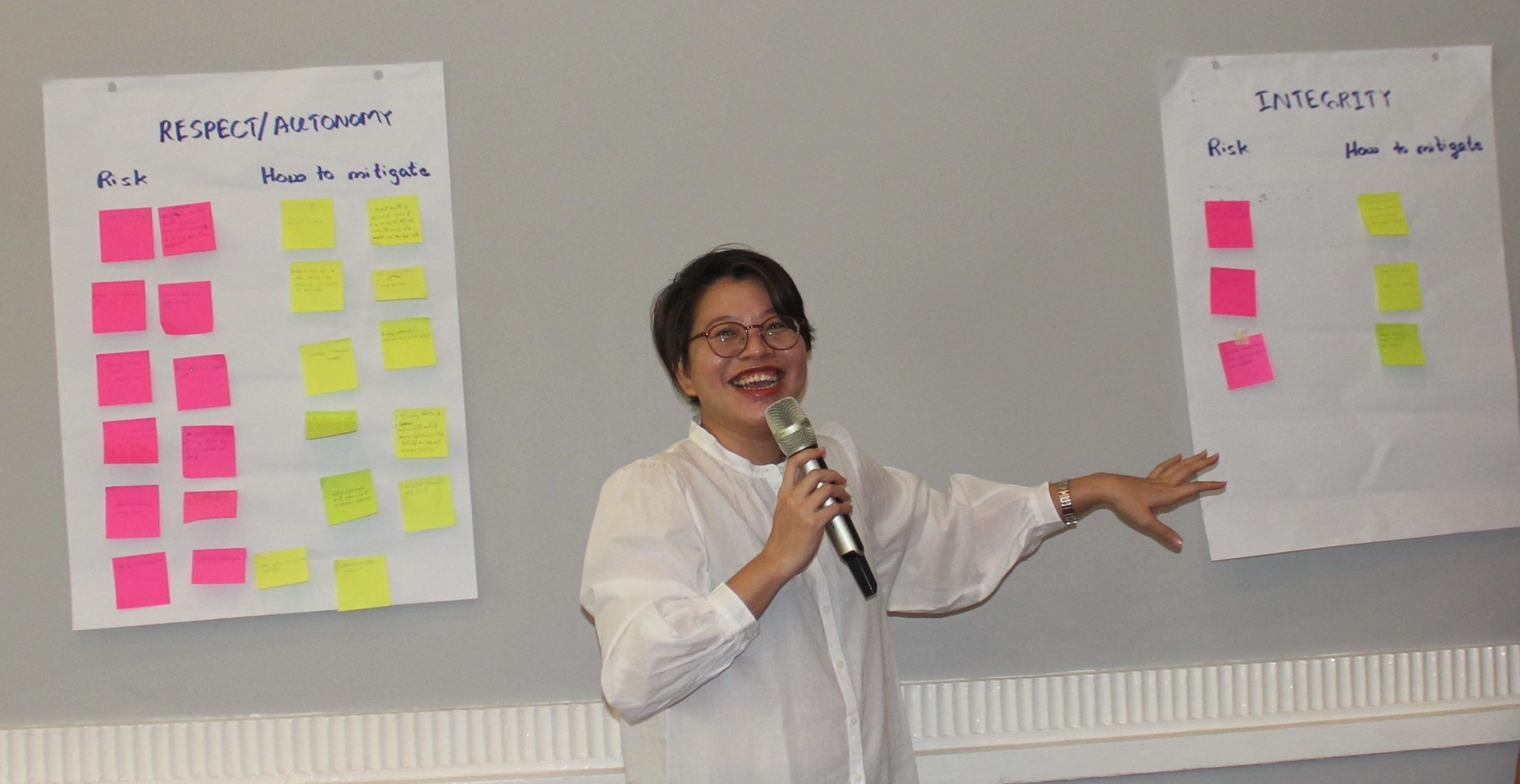 A smiling Burmese woman stands pointing to post-its on a wall while speaking into a microphone