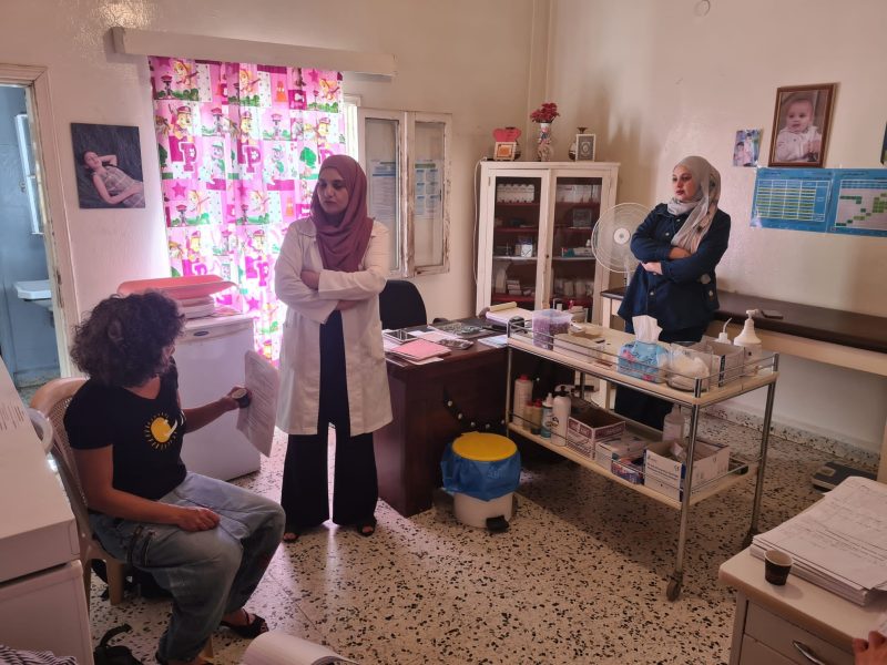 Two Muslim women stand talking to a seated woman in a medical examination room with pink, kids curtains