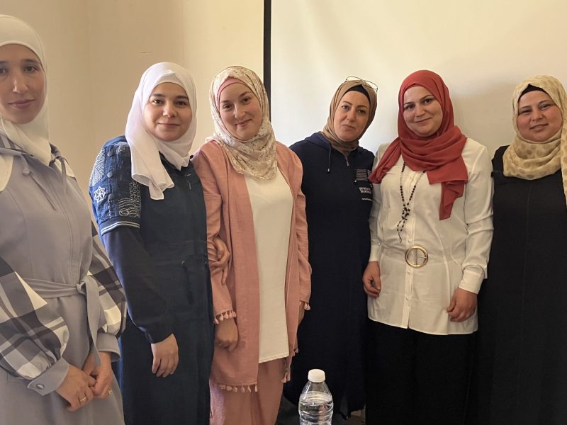 A group of six standing, smiling women wearing headscarves