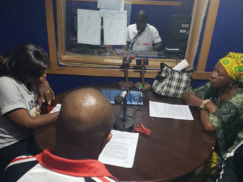 Three African people sit around a table - there are microphones and sheets of paper in front of them - while a man sits in a booth on the other side of some glass - he has a mixing desk in front of him