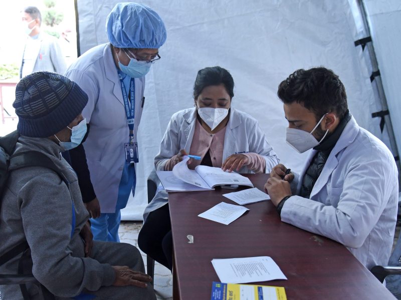 Three masked medics doing paperwork around a table while a masked man in grey warm clothes looks on