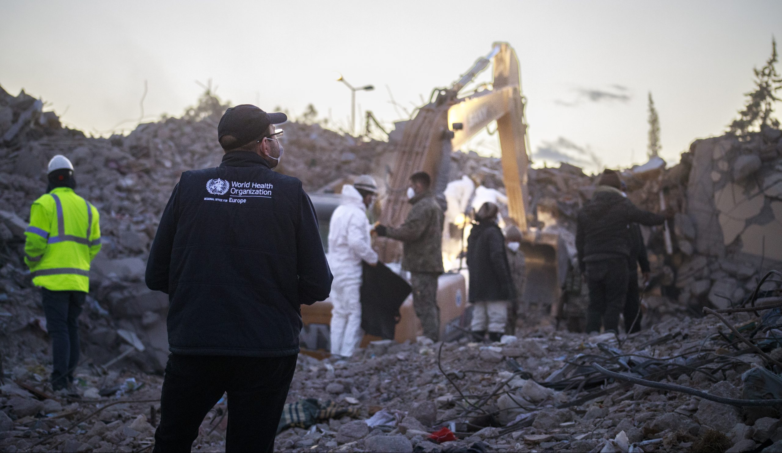 A man in a black WHO jacket stands looking as rescue people with a digger excavate a collapsed building