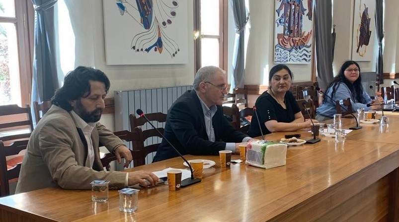 Two men and two women sit at a long wooden table in a room with art on the wall behind
