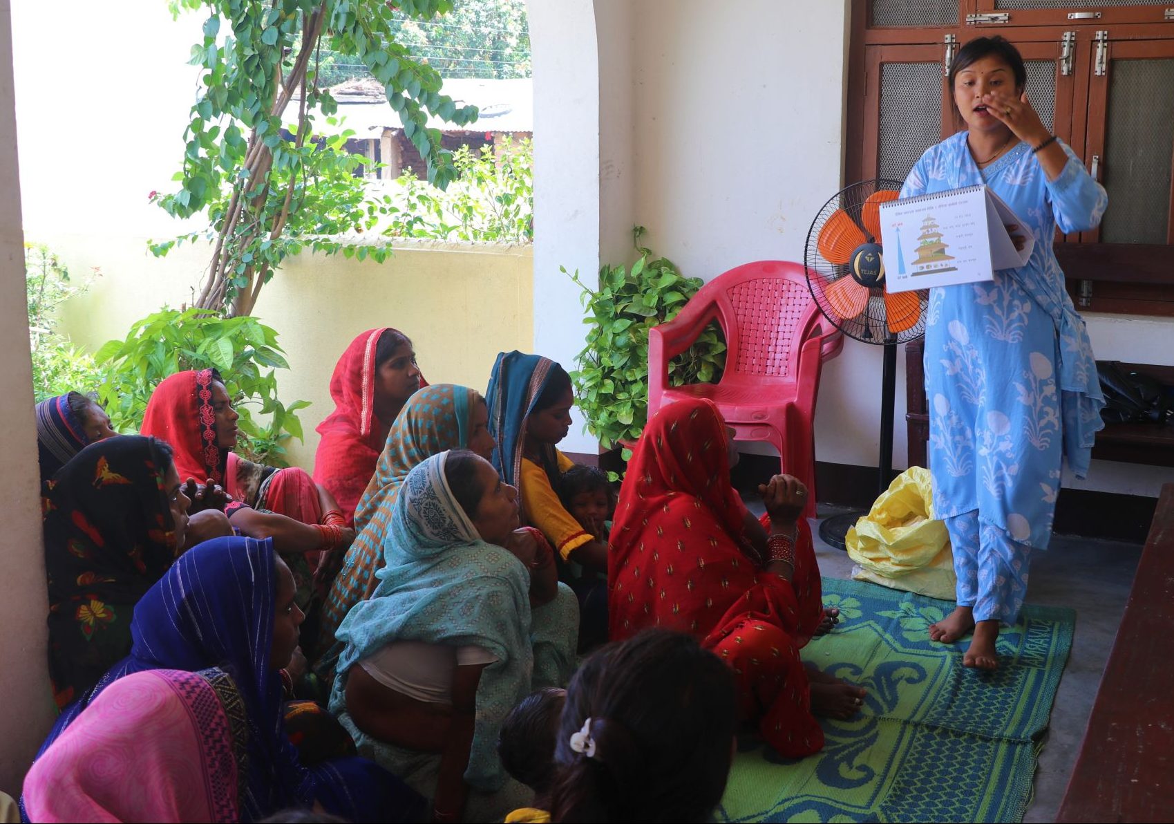 A female Community Health Volunteer in a blue sari stands in front of a group of seated women