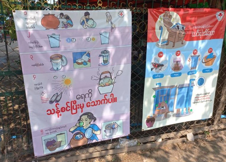 Two large posters attached to an outdoor railing. Both show simple illustrations of people using water and cooking