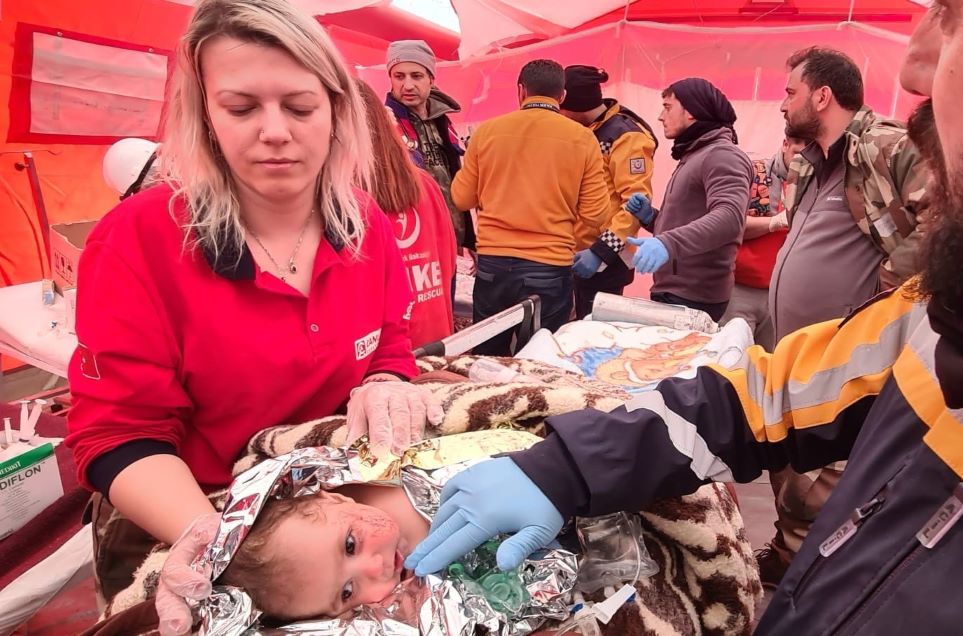 A female emergency worker cares for a baby wrapped in foil with scratches on their face. They are with other emergency health workers in an orange tent. Plus the title 'Strengthening health workforce emergency preparedness for future shocks: Lessons from the 2023 earthquake in Türkiye'