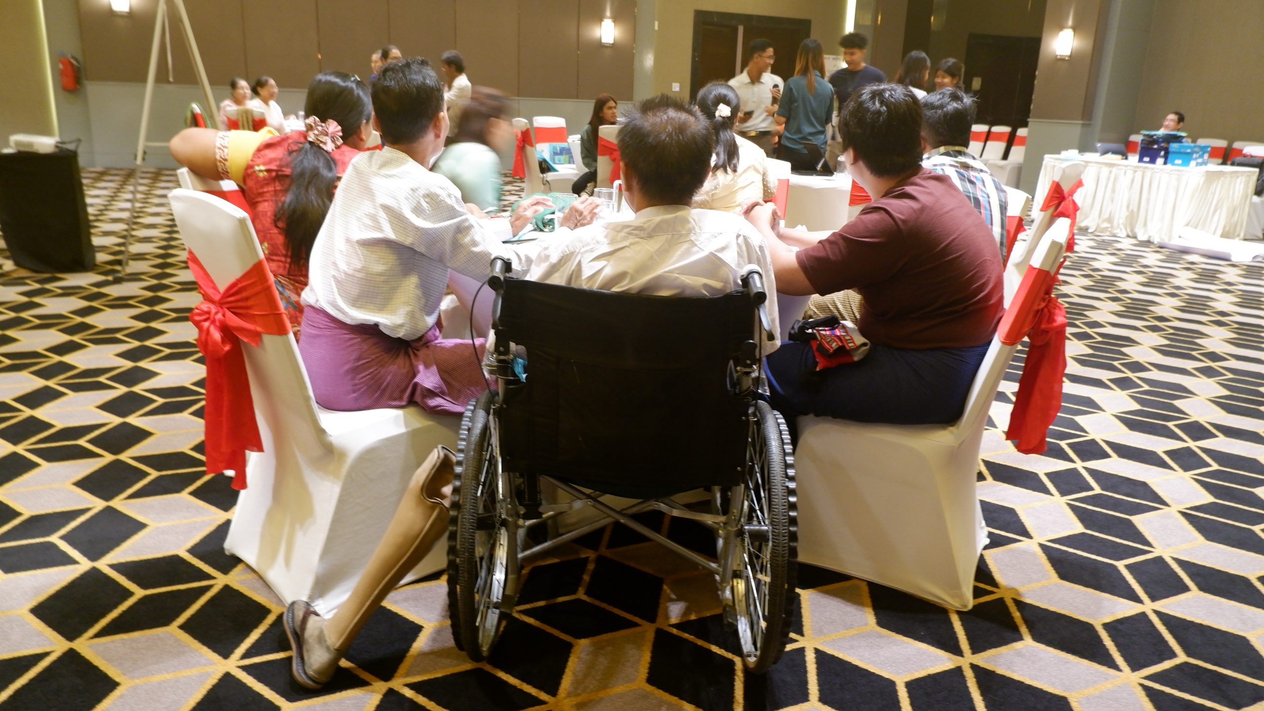 a large meeting room with Burmese people sitting around a large table. The person sitting closest to the camera is sitting in a wheelchair. All have their backs to the viewer.