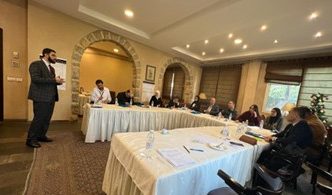 A Syrian man in a dark suit stands in front of a room of seated men and women who are listening to him talk