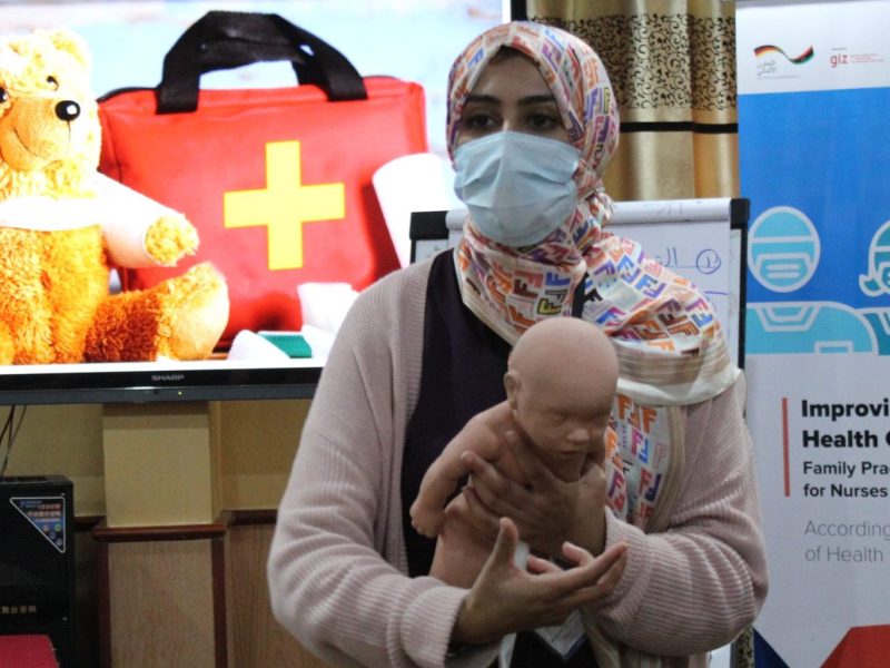 A masked Libyan women stands holding a plastic baby, demonstrating a medical technique. She's in front of a poster on 'Improving primary health care in Libya'