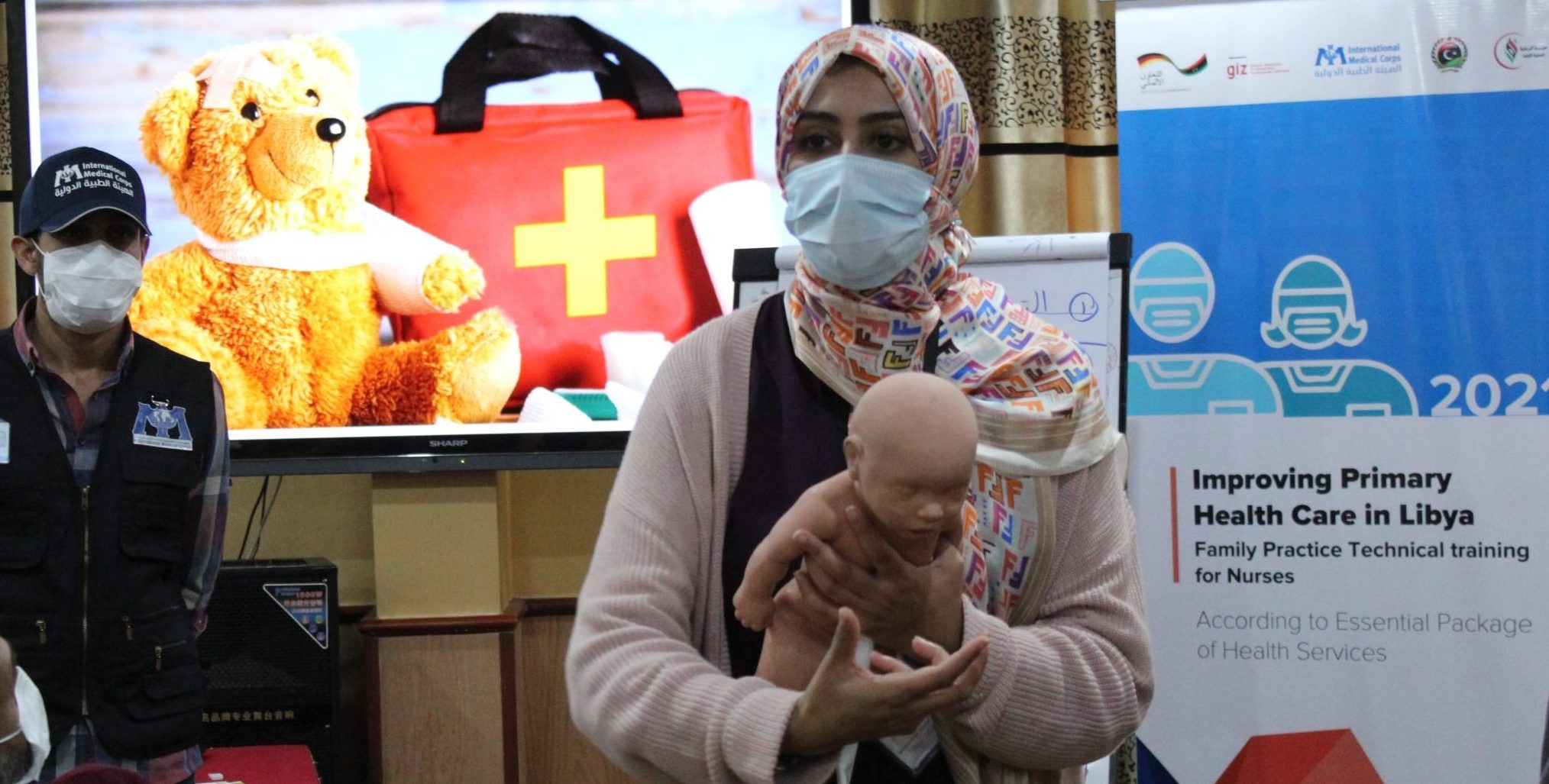 A masked Libyan women stands holding a plastic baby, demonstrating a medical technique. She's in front of a poster on 'Improving primary health care in Libya'