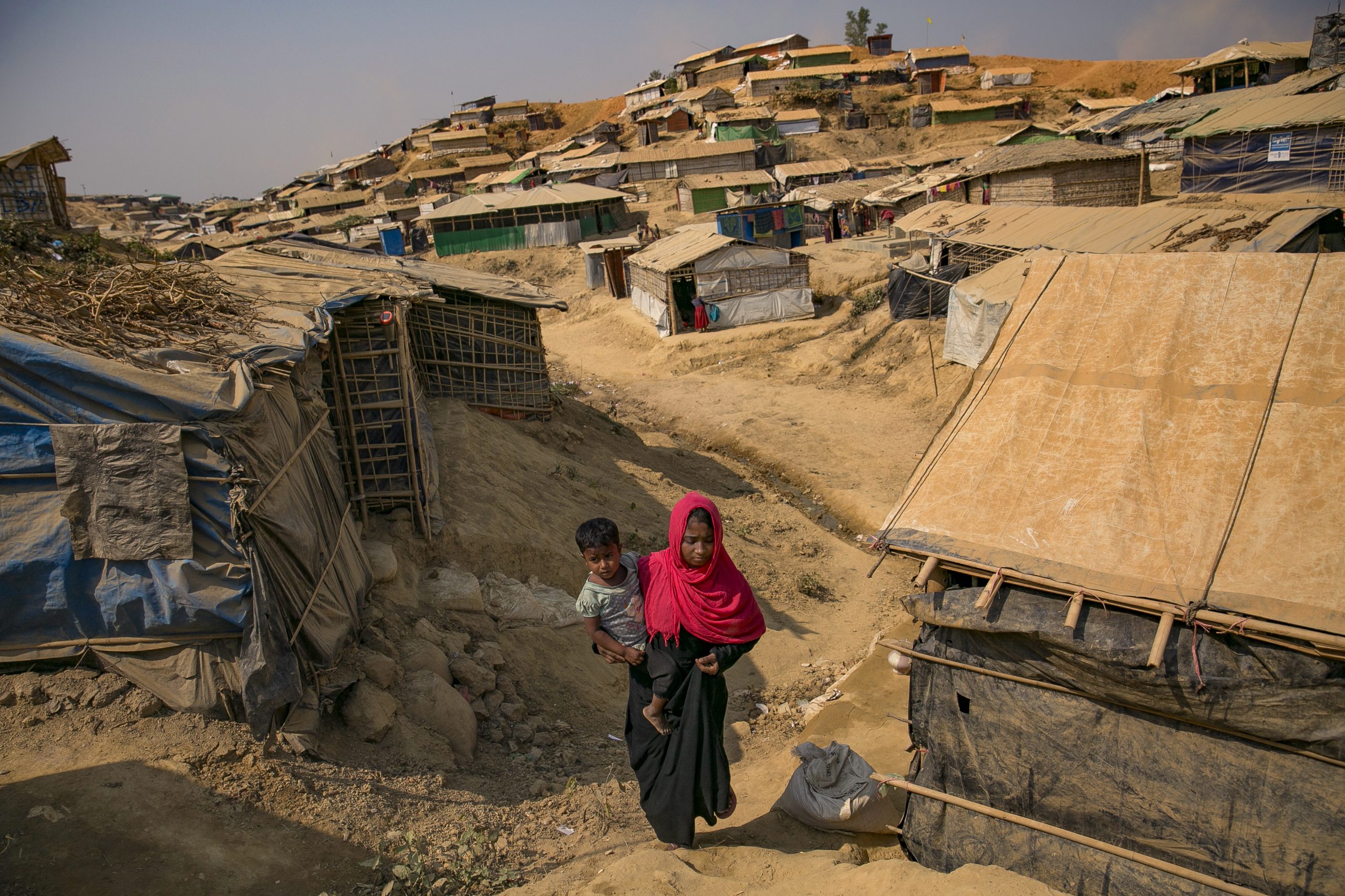 A young Rohingya woman carries a baby through a sandy refugee camp - they are surrounded by tents